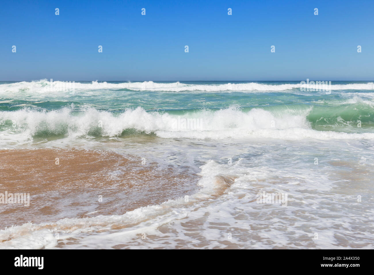 Low angle frontal view of small foamy ocean waves crashing on a sandy beach on a sunny day. Stock Photo