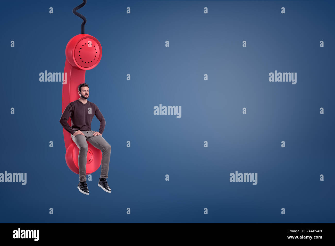 A casual looking bearded man sits on a giant red retro phone receiver hanging from a cord. Stock Photo