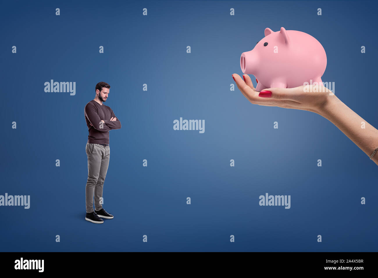 A small sad bearded man stands near a large female hand holding a piggy bank. Stock Photo