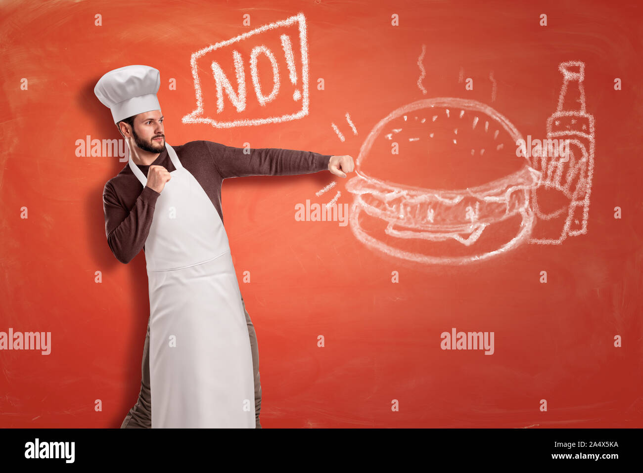 A young handsome chef saying 'No' and hitting with clenched fists a chalk drawing of a hamburger, French fries and a ketchup bottle. Stock Photo