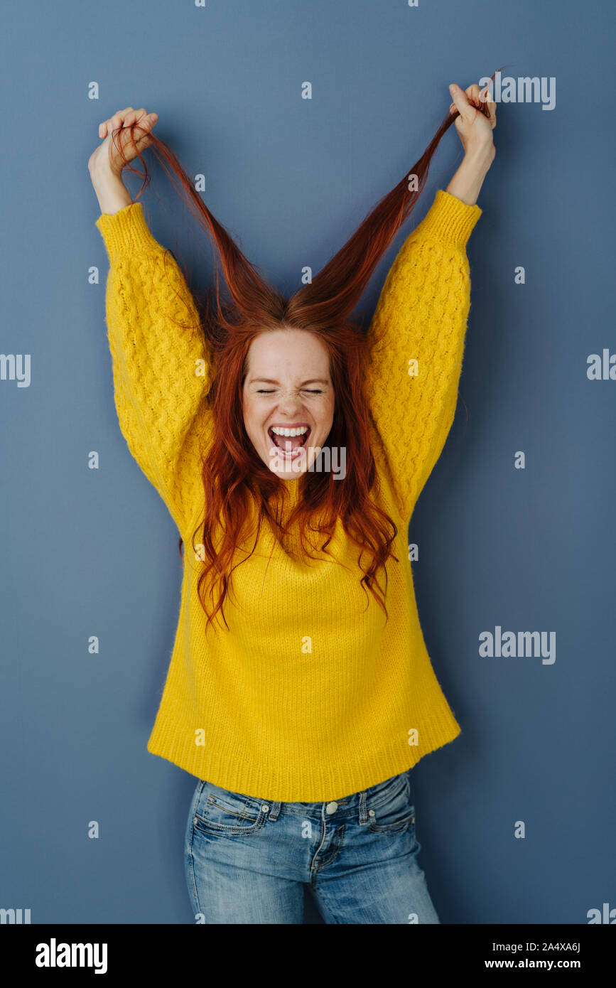 Elated casual young woman cheering and pulling up her long red hair in her hands over a blue studio background Stock Photo