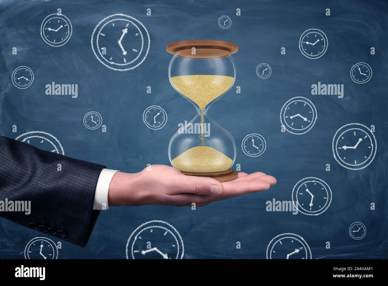 A businessmans hand turned palm up and holds a large functioning hourglass on blue chalkboard background. Stock Photo