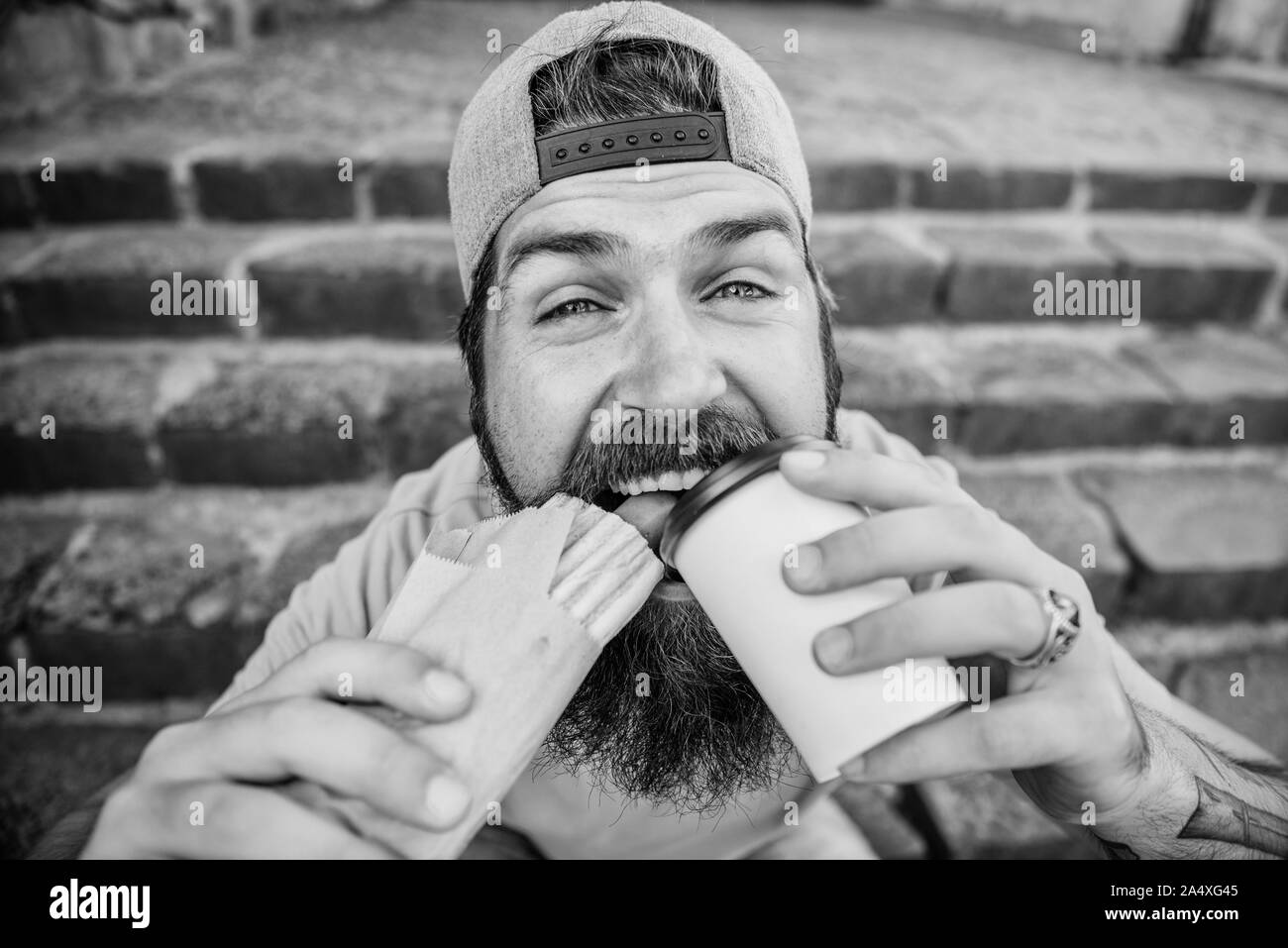 Street food concept. Man bearded eat tasty sausage and drink paper cup. Urban lifestyle nutrition. Junk food. Carefree hipster eat junk food while sit stairs. Guy eating hot dog. Snack for good mood. Stock Photo