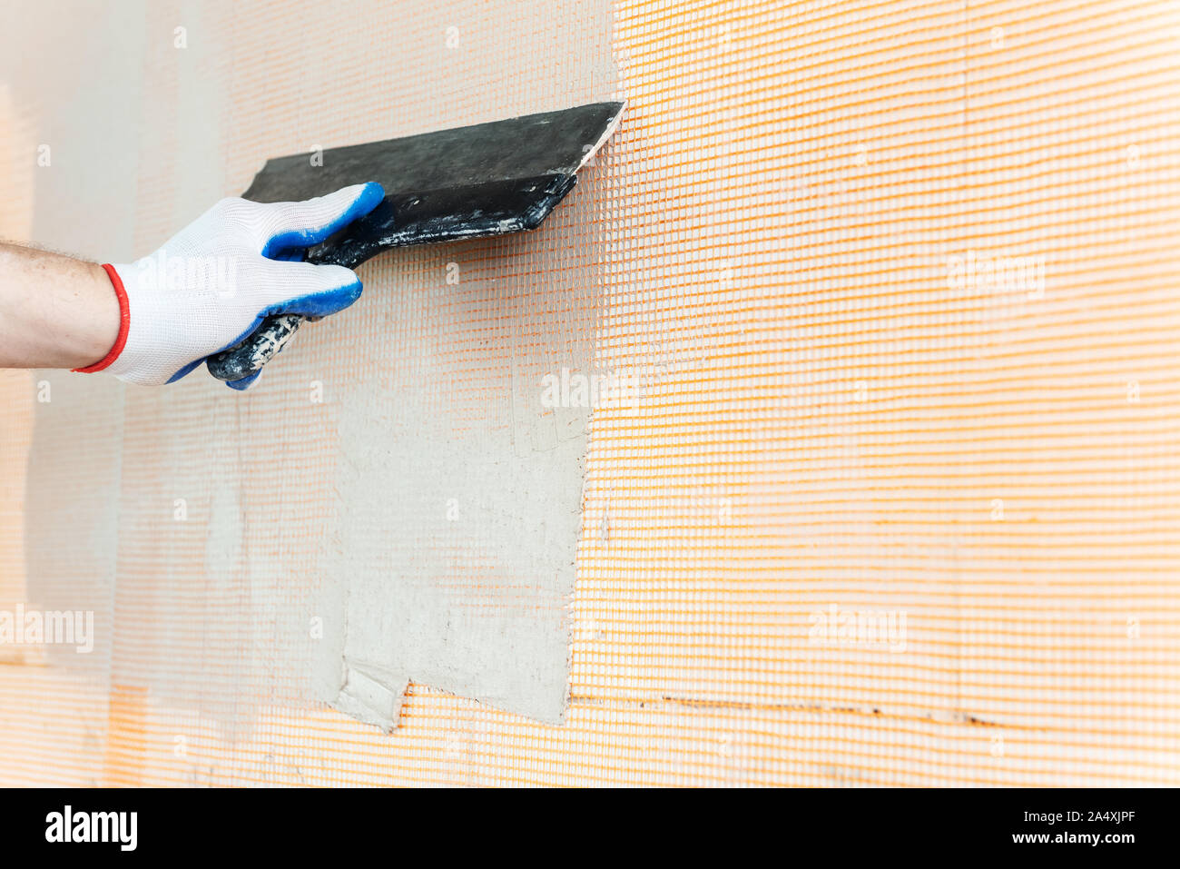 A worker is applying putty on a fiberglass mesh on the wall. He is using a wide spatula. Stock Photo