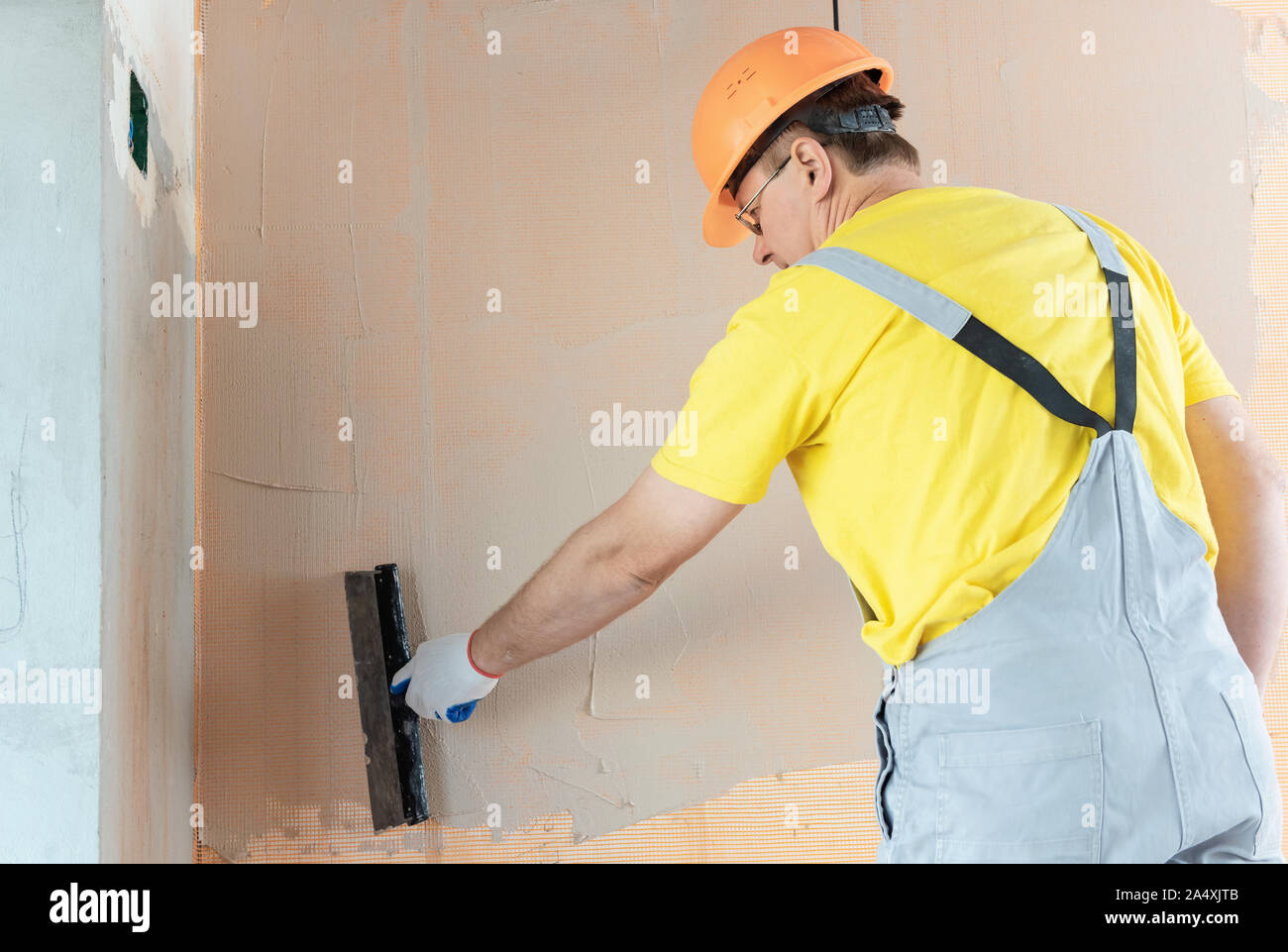 The worker is applying putty on a fiberglass mesh on the wall. He is using a wide spatula. Stock Photo