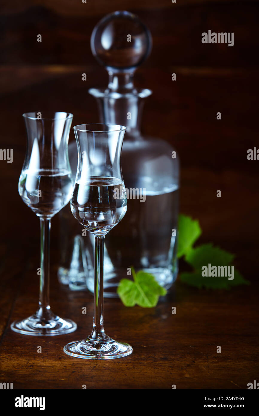 Two high glasses of grappa bianca and decanter with round stopper in background in contrast light still life Stock Photo