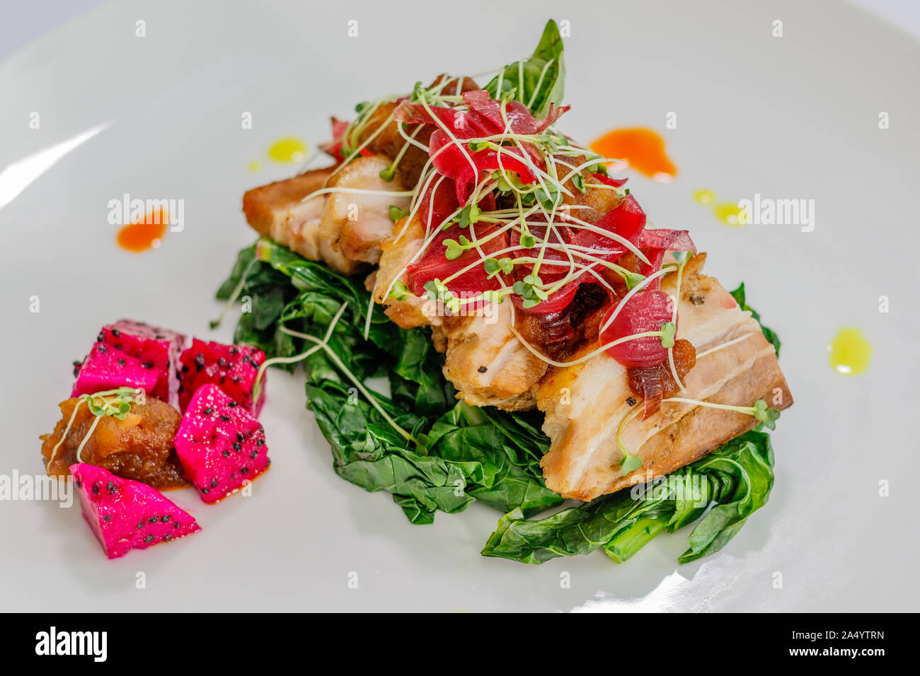 White ceramic plate with pork belly with Thai salad decorated with onion colored with beetroot sauce, bean sprouts and dragon fruit. Side view. Stock Photo