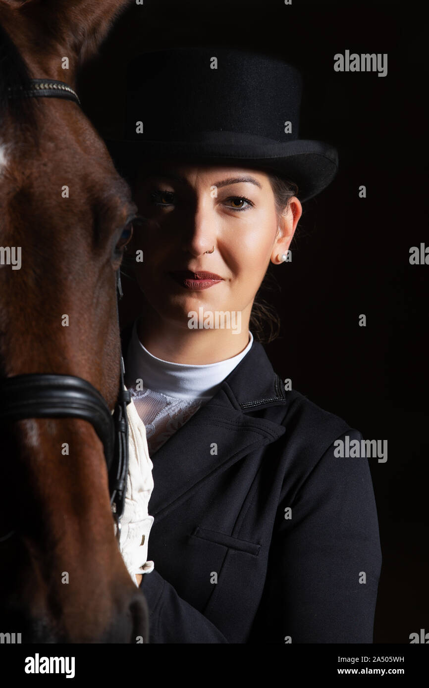 Young woman in tails in portraits next to her horse. She is holding her head next to the one of the horse and is looking into the camera. Stock Photo