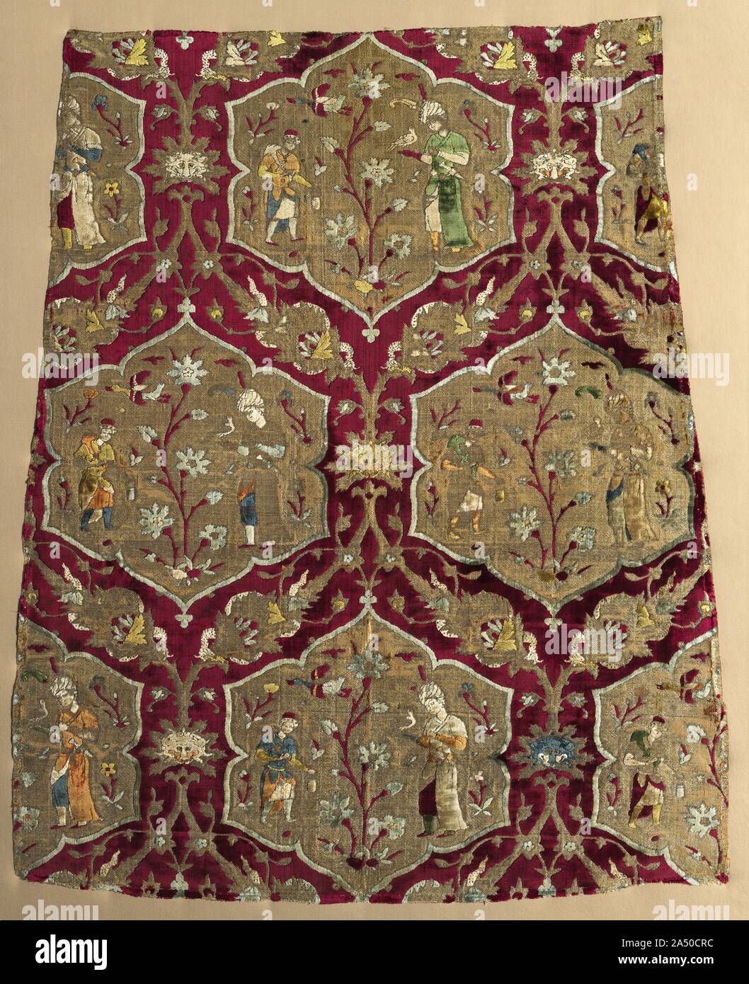 Brocaded velvet with falconer and attendant in animated lattice, from a robe, mid 1500s. This signature Safavid velvet displaying a falconry scene is celebrated for its refined beauty, meticulous draftsmanship, and exemplary technique with eight colours of velvet pile. A falconer and attendant flank a tall blossoming plant on a golden ground of lobed medallions. Animated foliate vines display leaves bearing lion&#x2019;s masks and dragons coiled around larger leaves on the rich crimson velvet ground. Instead of two or three colours of velvet pile, ingenious Iranian weavers wove velvets with as Stock Photo