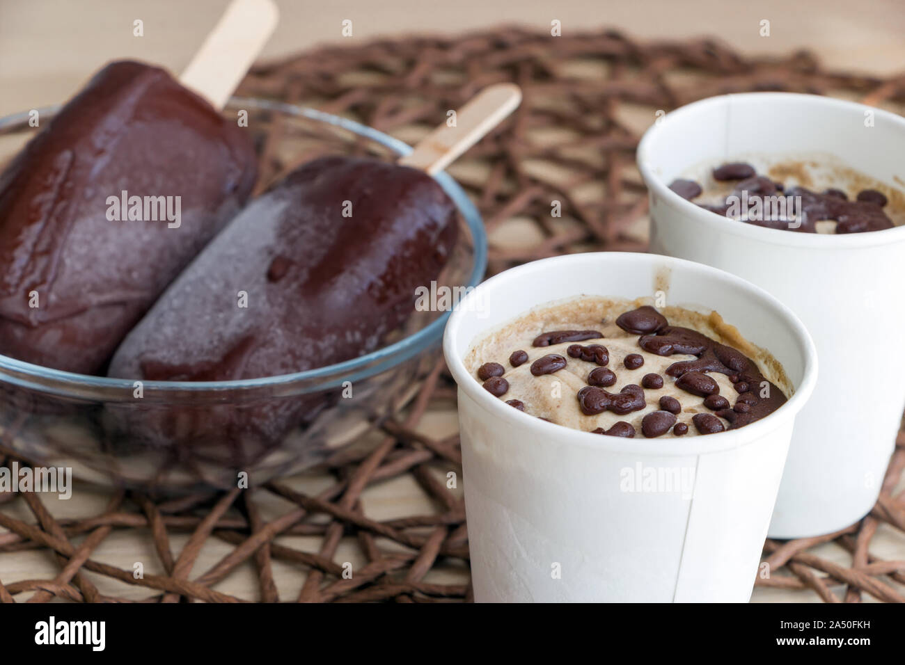 Raw banana ice cream with coconut cream, coated with natural chocolate, in a white paper cup, and chocolate popsicles on sticks in glass bowl. Healthy Stock Photo