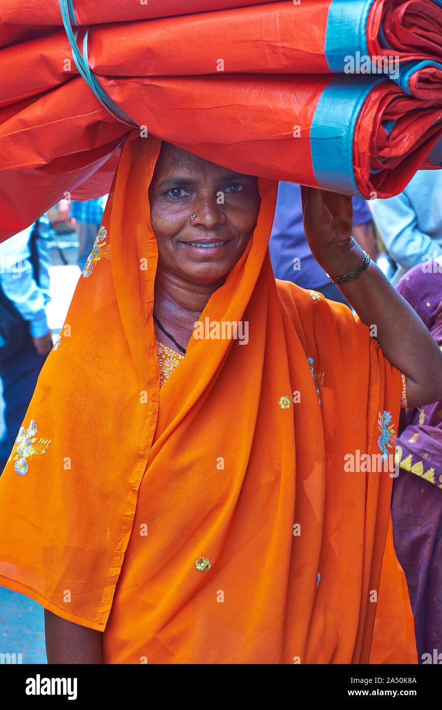 A woman dressed in an orange sari carries red cloth on her head, purchased to earn extra income for the family; at Manish Market, Mumbai, India Stock Photo