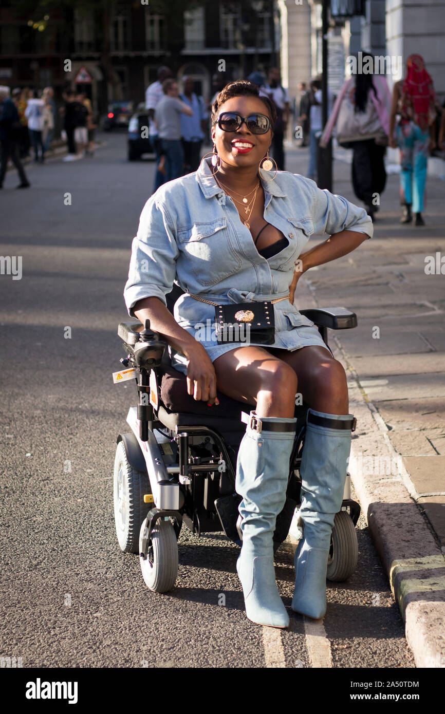 LONDON, UK- SEPTEMBER 13 2019: People on the street during the London Fashion Week. Girl in a denim shirt and a short leather skirt sits in a wheelcha Stock Photo