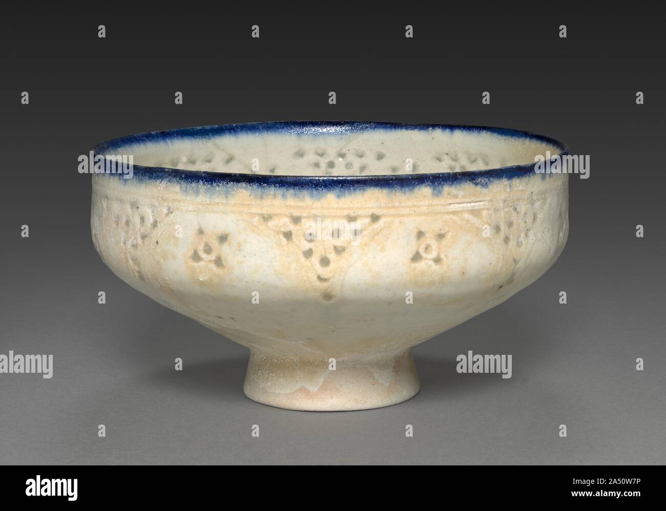 Footed Bowl, late 1100s-early 1200s. Iranian potters created transparency by piercing and glazing frit-body walls. Here, triangles and rosettes decorate incised scallops beneath the blue rim. The footed shape is derived from Iranian metal bowls. Stock Photo
