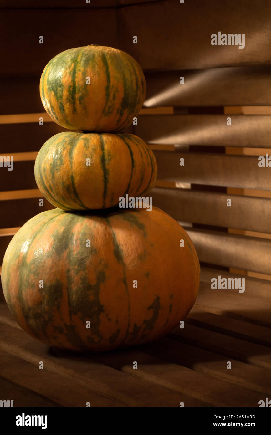 A pyramid of three pumpkins on a wooden surface against the background of walls of wooden slats through which light breaks through with beautiful rays Stock Photo