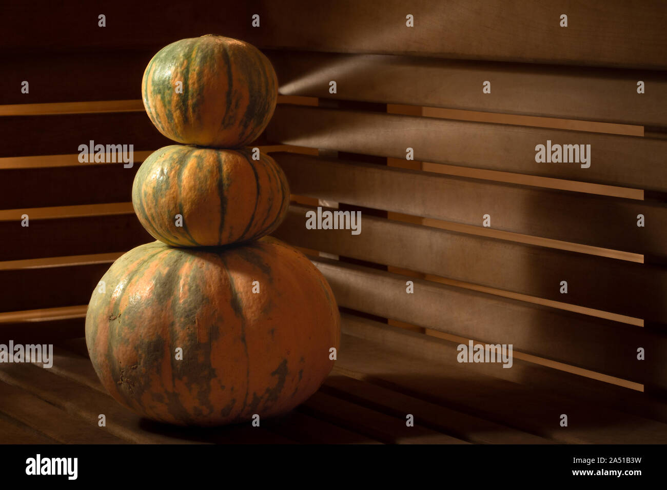 A pyramid of three pumpkins on a wooden surface against the background of walls of wooden slats through which light breaks through with beautiful rays Stock Photo