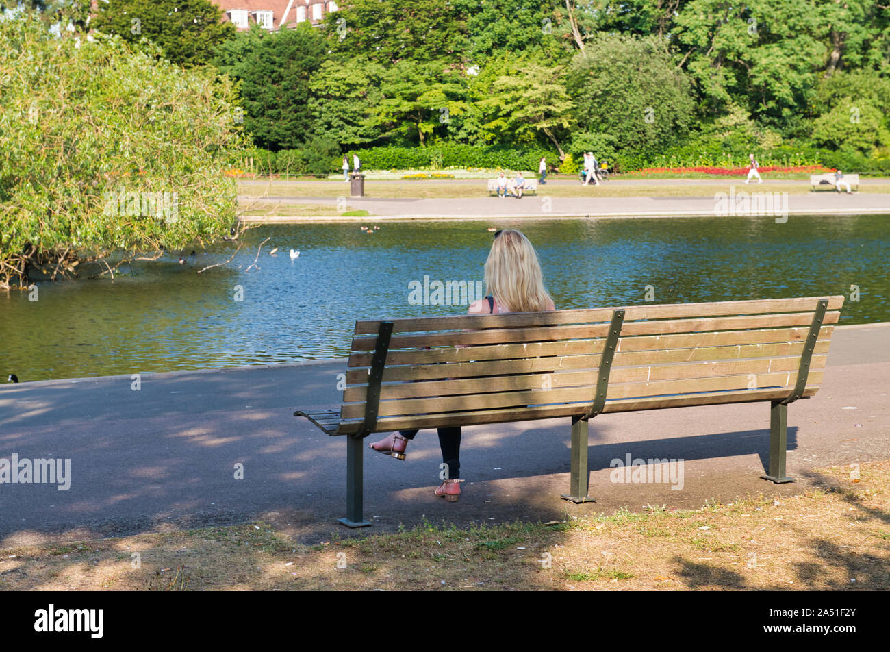 Back view of blonde woman seated on a bench along the lake. Stock Photo