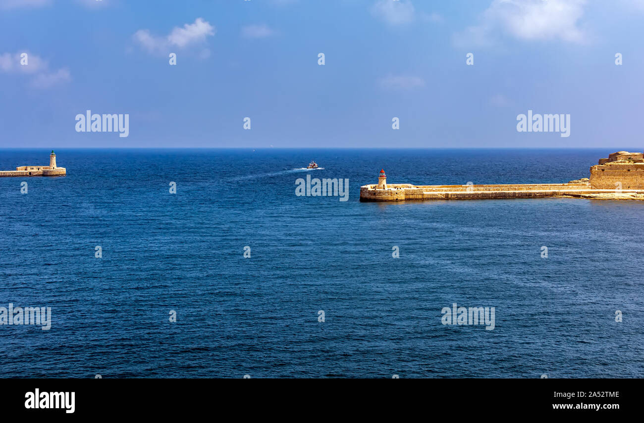 Boat going in turquoise sea water between Valletta breakwater with lighthouse and Ricasoli Breakwater Light at Grand Harbour East Breakwater. Stock Photo