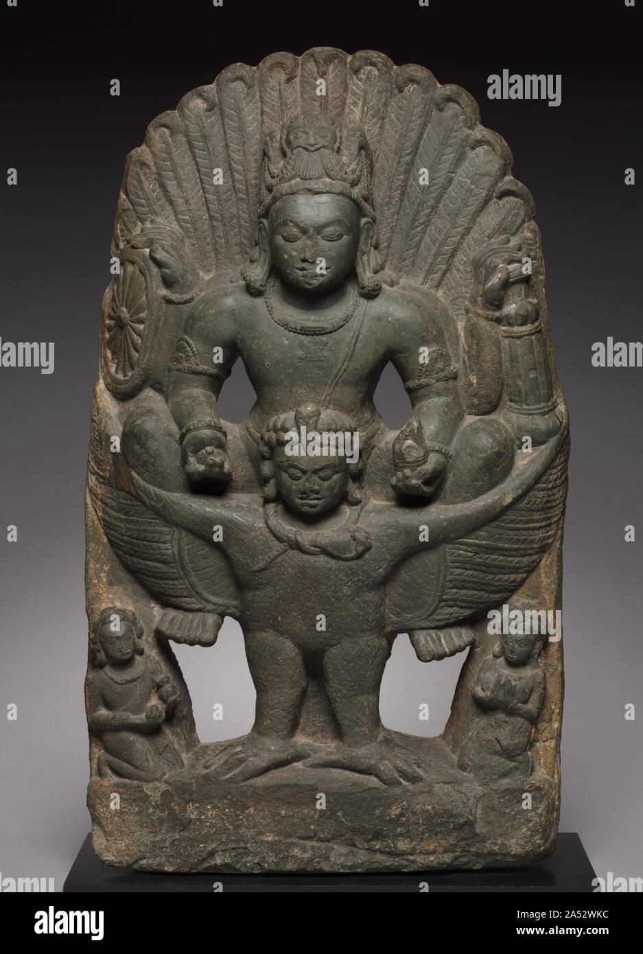 Vishnu Riding on Garuda, 500s-600s. In a rare and powerful work from this period, Vishnu straddles his vehicle, the man-eagle called Garuda, here shown with a human head, wings outstretched, and tail feathers expanding like rays of light behind the head of the god. Garuda is the enemy of serpents, and a subjugated serpent is tied around his neck. His determined expression lends confidence that he will do anything in his power to support Vishnu in his accomplishments. Tiny worshippers kneel in veneration before the imposing figures.   Each of Vishnu's hands holds an implement: discus, mace, con Stock Photo