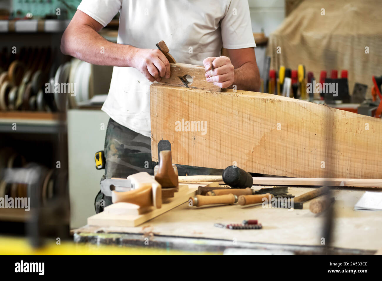 Carpenter planing a wooden block with a manual planer smoothing or reducing the surface with assorted tools in the foreground in a woodworking worksho Stock Photo