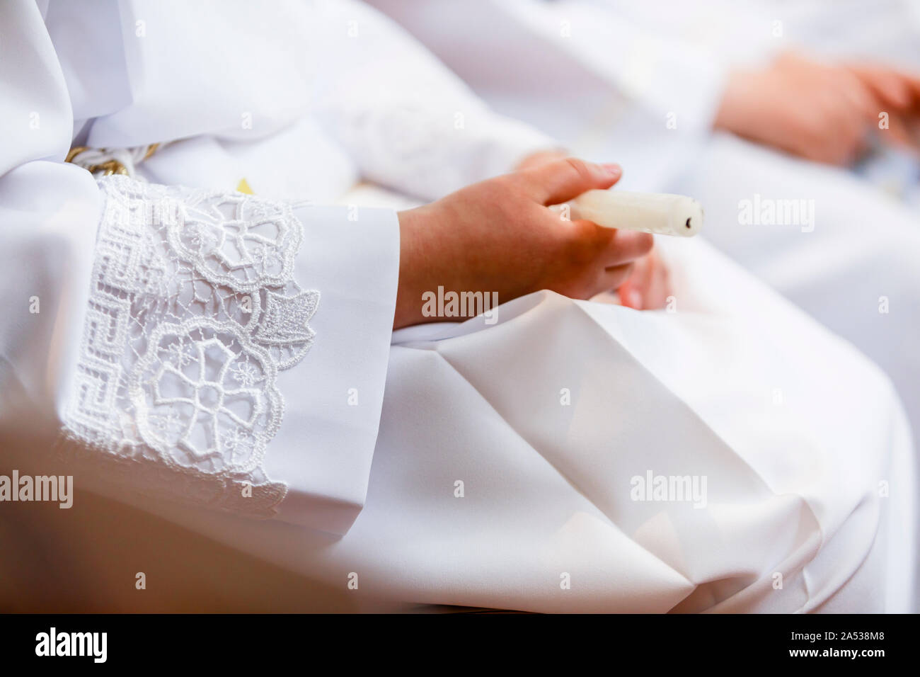Close-up of a white lace ceremonial robe and a hand holding a handmade candle at a first communion catholic ceremony in church. Light and airy, with c Stock Photo