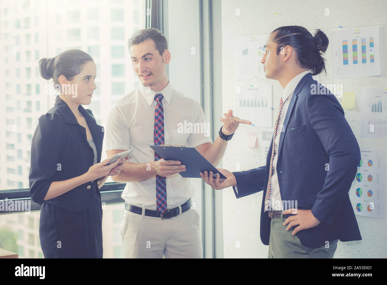 Three businesspeople standing in modern office looking file document and talking in meeting room. Stock Photo