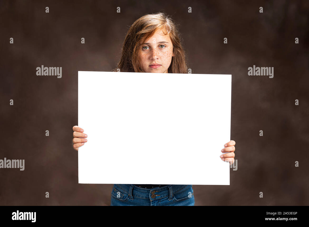 Horizontal studio shot of a pre-teen girl with beautiful eyes holding a blank white sign.  She has a serious expression on her face.  Brown background Stock Photo