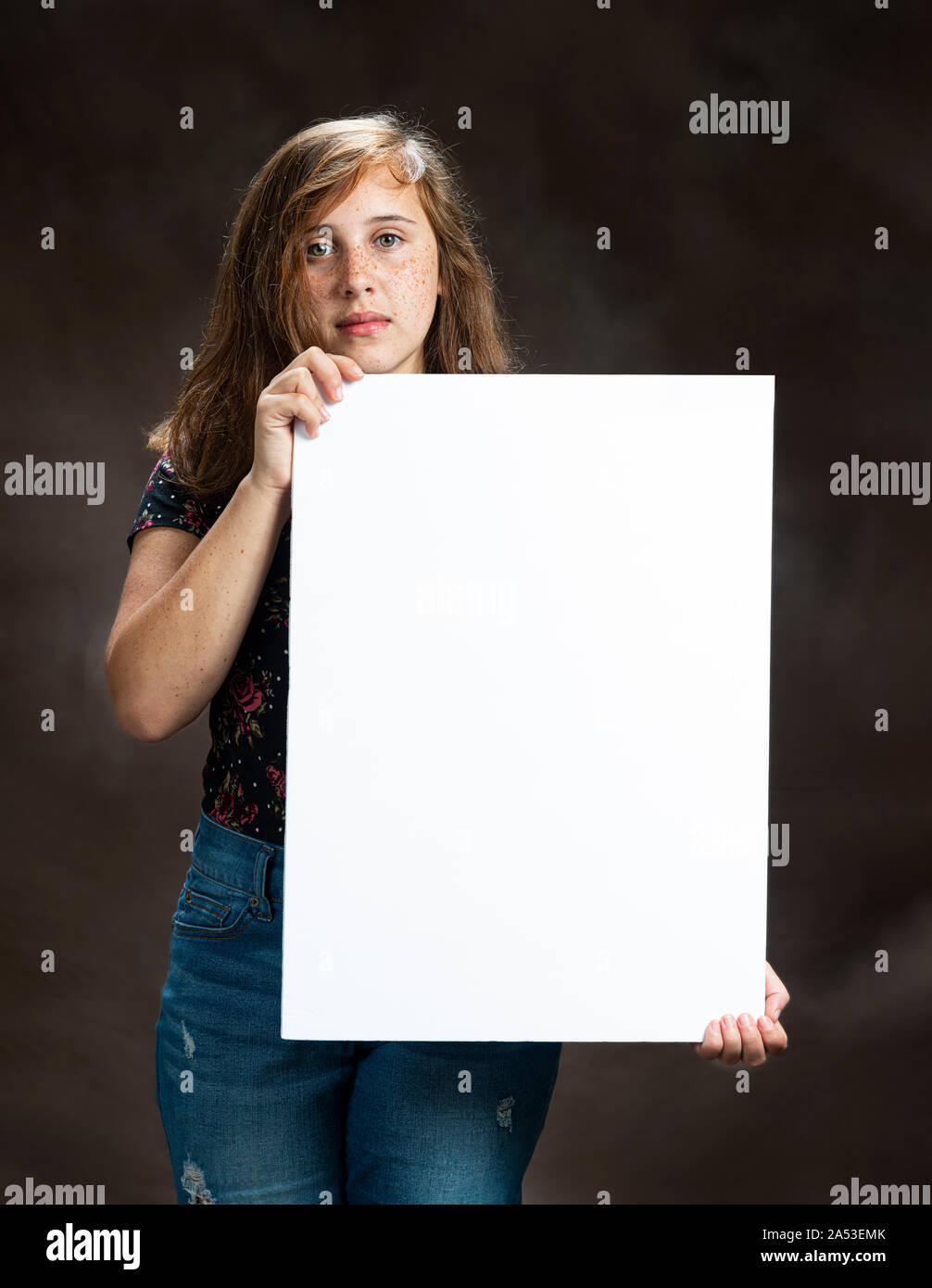 Vertical studio shot of a serious looking pre-teen girl holding a blank vertical white sign.  Brown background. Stock Photo