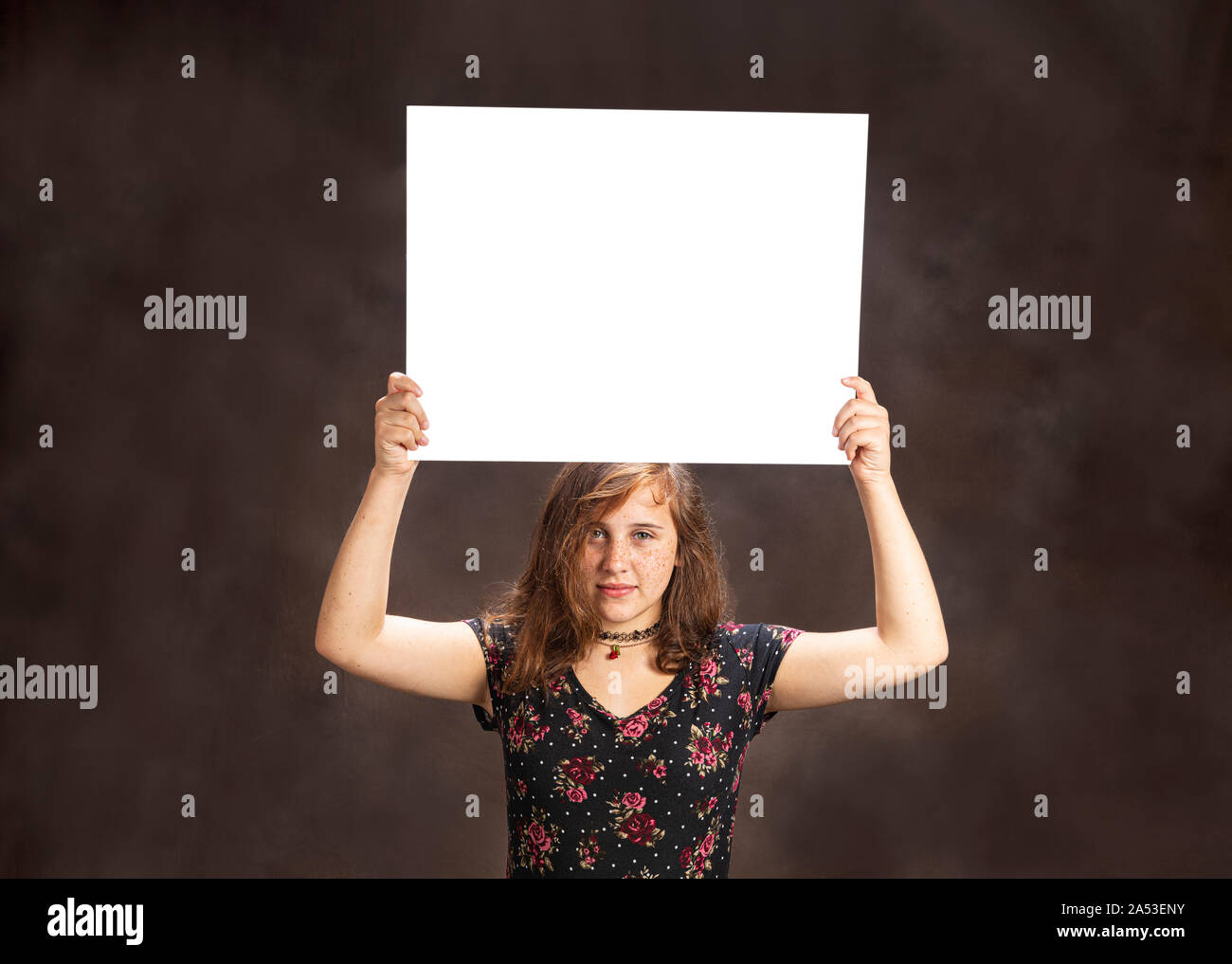 Horizontal studio shot of a serious looking pre-teen girl with freckles holding a blank white sign on her head.  Brown background.  Copy space. Stock Photo