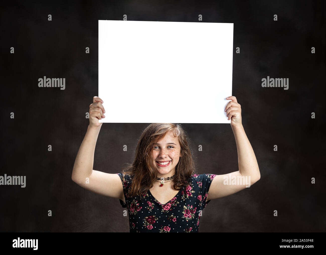 Horizontal studio shot of a happy pre-teen girl with freckles holding a blank white sign on her head on a brown background. Stock Photo