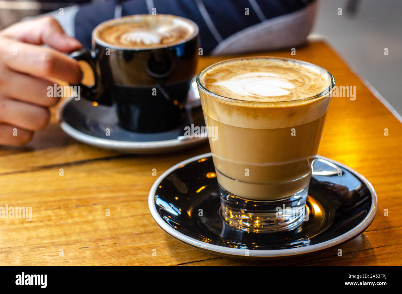 Close up of a cup of cafe latte with a blurry background of a man's hand holding his coffee on the opposite side of the table. Stock Photo