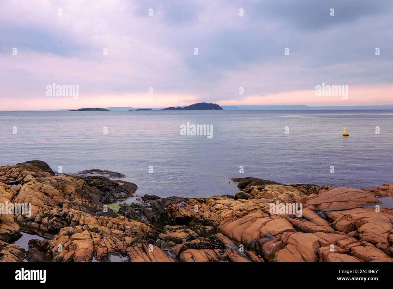 View on insels on Oslo Fjord, calm water, overcast sky. Landscape with orange rocks on the beach. Norwegian coastline in summer. Nesodden Norway. Stock Photo