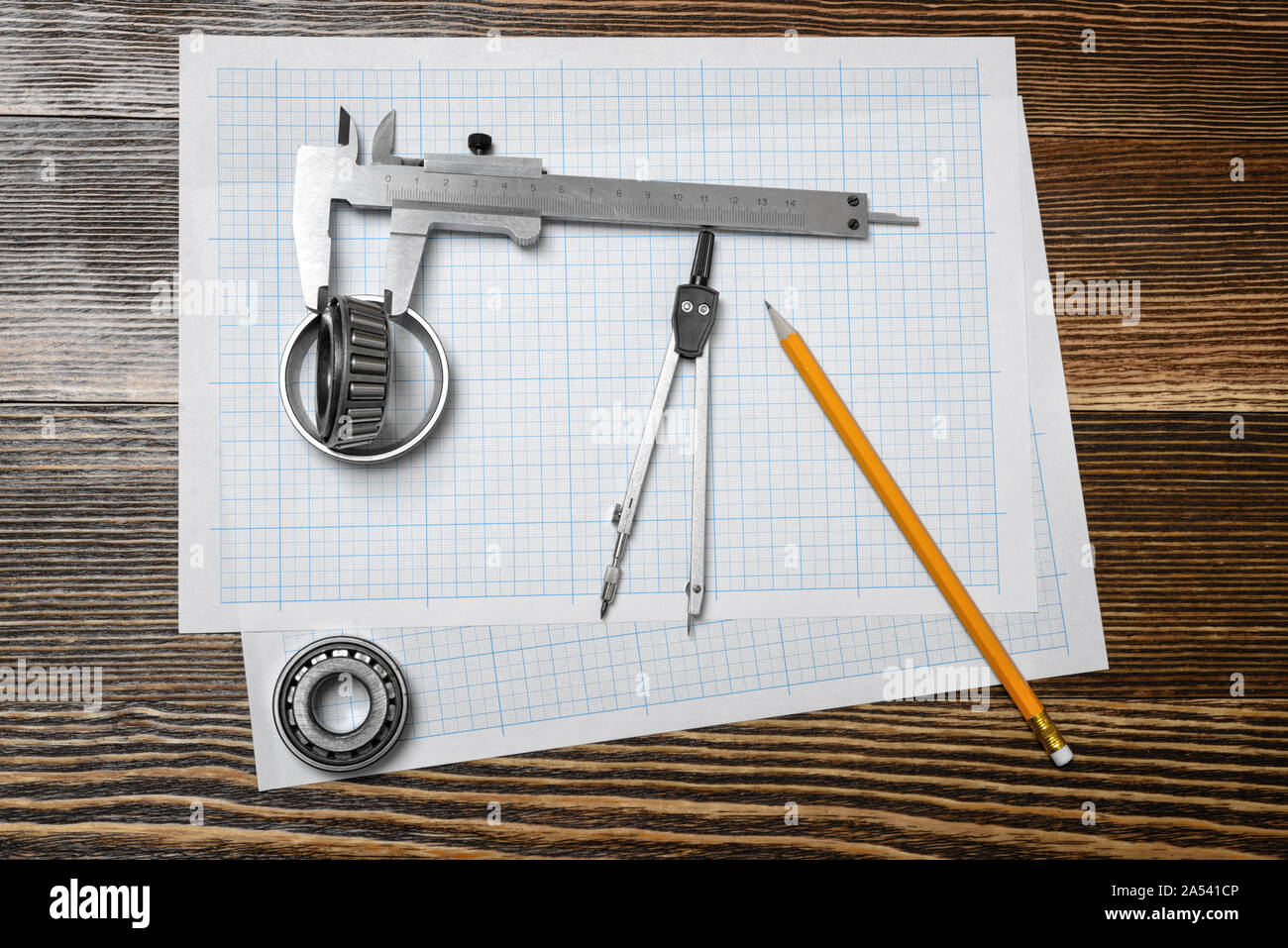 A vernier caliper holding a bearing, a pencil and a pair of compasses lying over drafting paper on wood background. Stock Photo