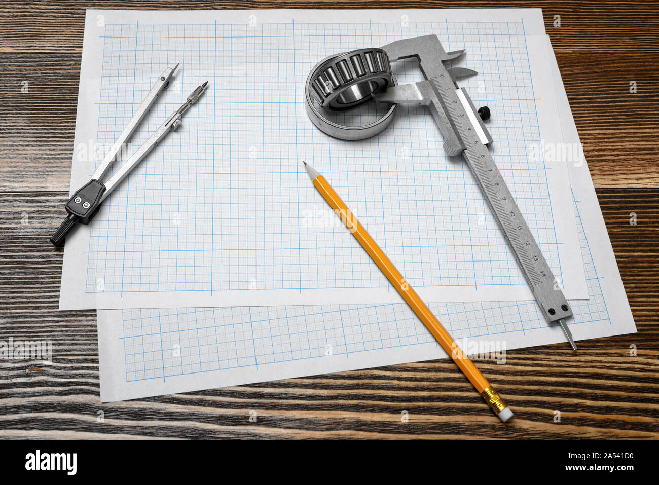 A vernier caliper holding a bearing, a pencil and a pair of compasses lying over drafting paper on wood background. Stock Photo