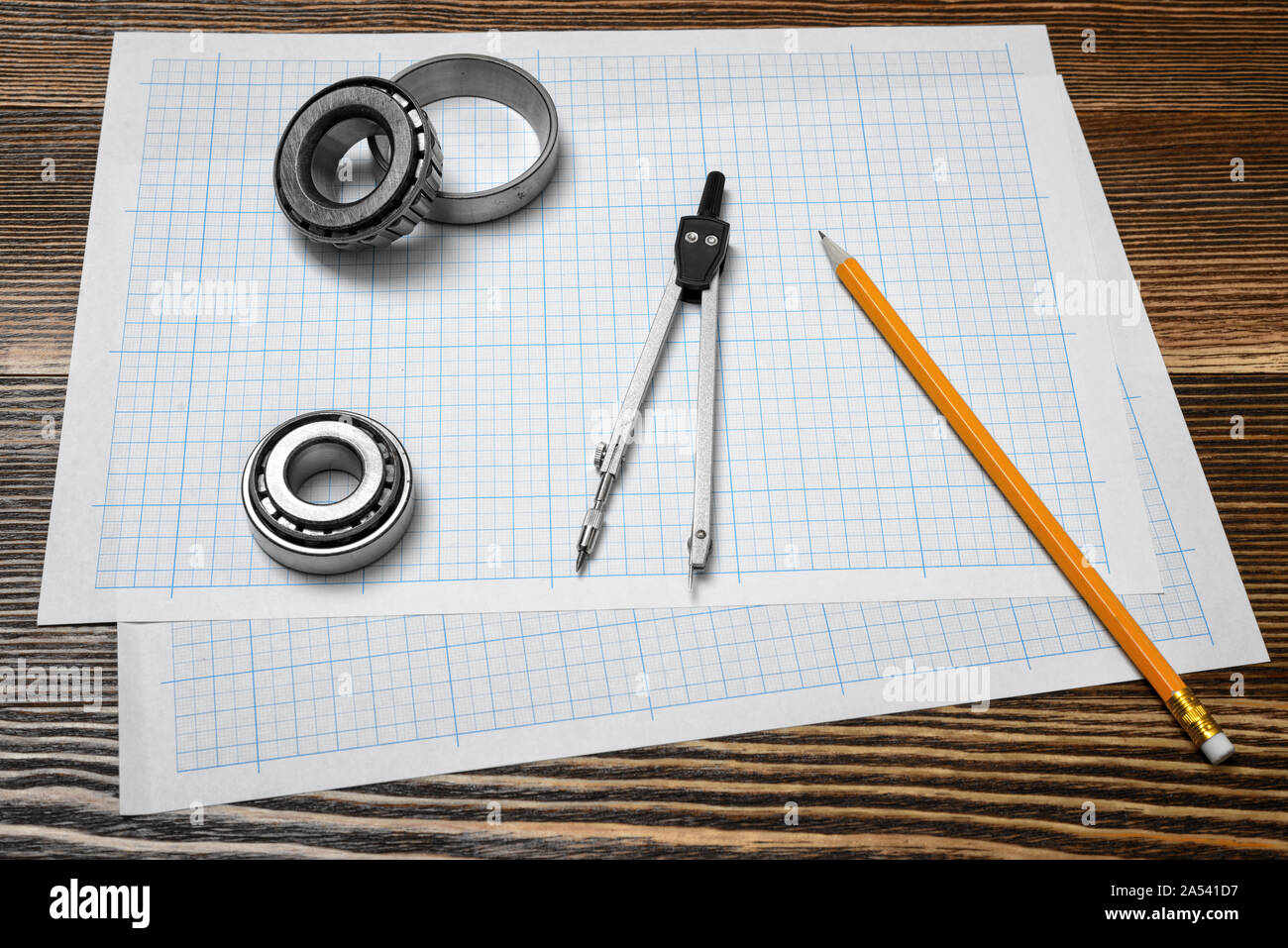 A vernier caliper holding a bearing, a pencil and a pair of compasses lying over drafting paper on wood background. Stock Photo