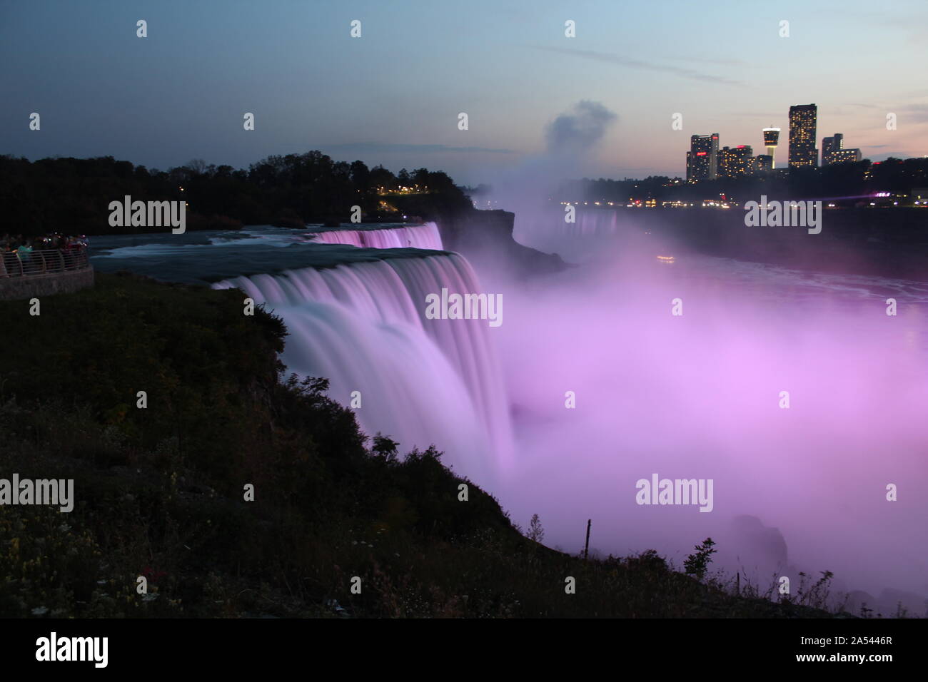Wide view of Niagara Falls illuminated pink in evening as seen from USA, with water flow all blurred, cityscape visible on Canada side Stock Photo