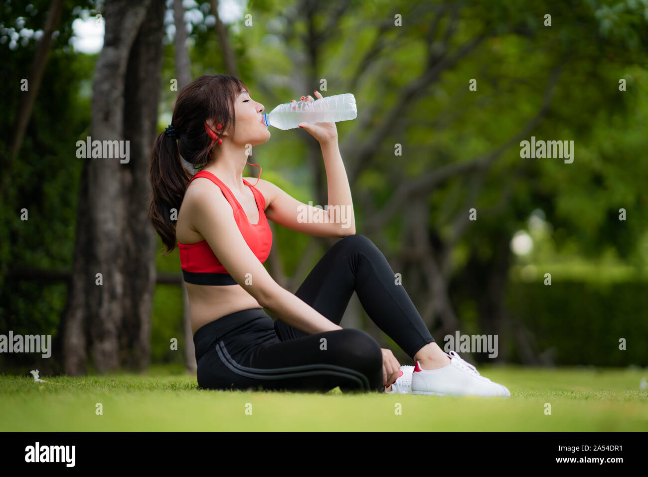 Asian beautiful woman sitting for relax after workout alone and drinking water from bottle in public park in village, happy and smile in morning durin Stock Photo