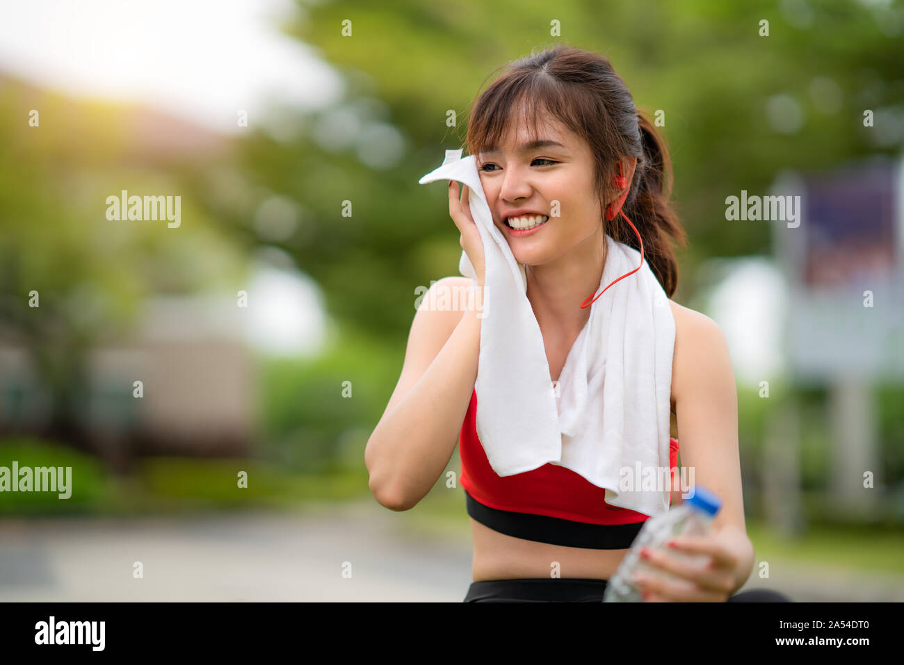 Asian beautiful woman sitting for relax after workout alone and holding water bottle in public park in village, happy and smile in morning during sunl Stock Photo