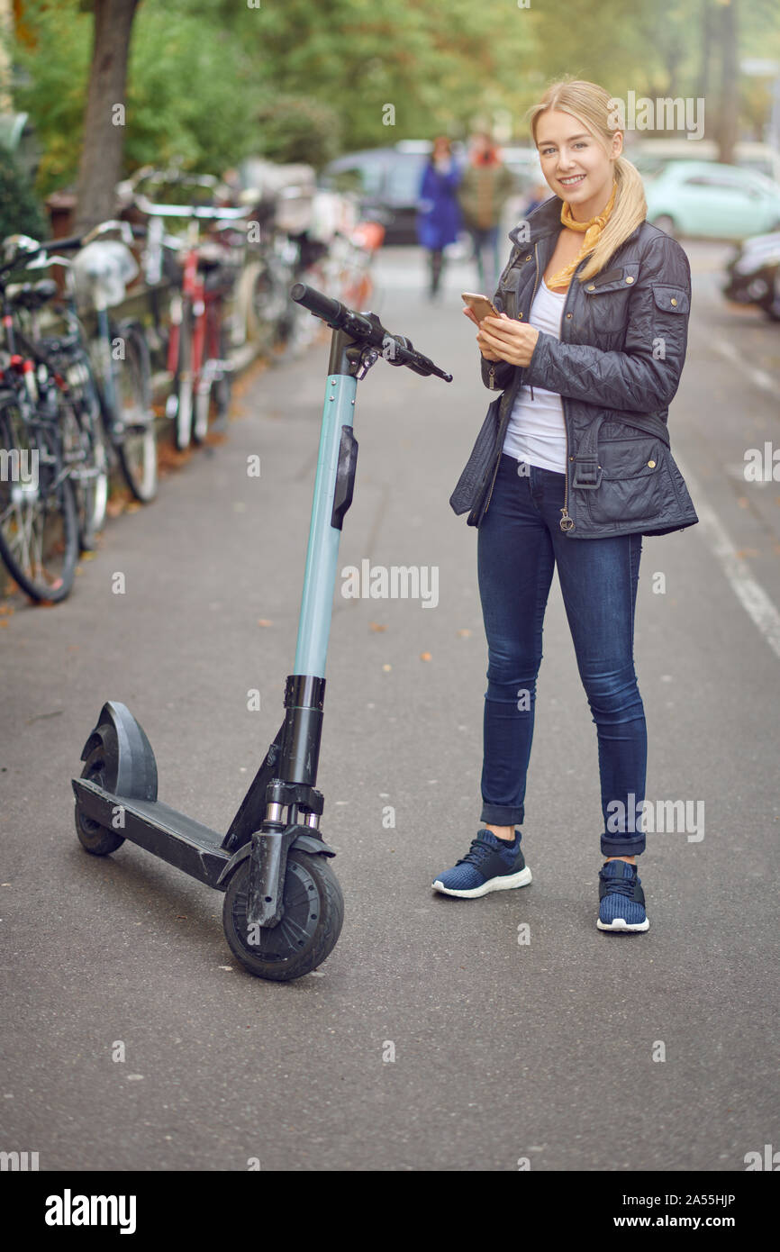 Young blond woman standing near electric kick scooter on city street or park, with bicycles parked on the side. Holding phone in her hands and smiling Stock Photo