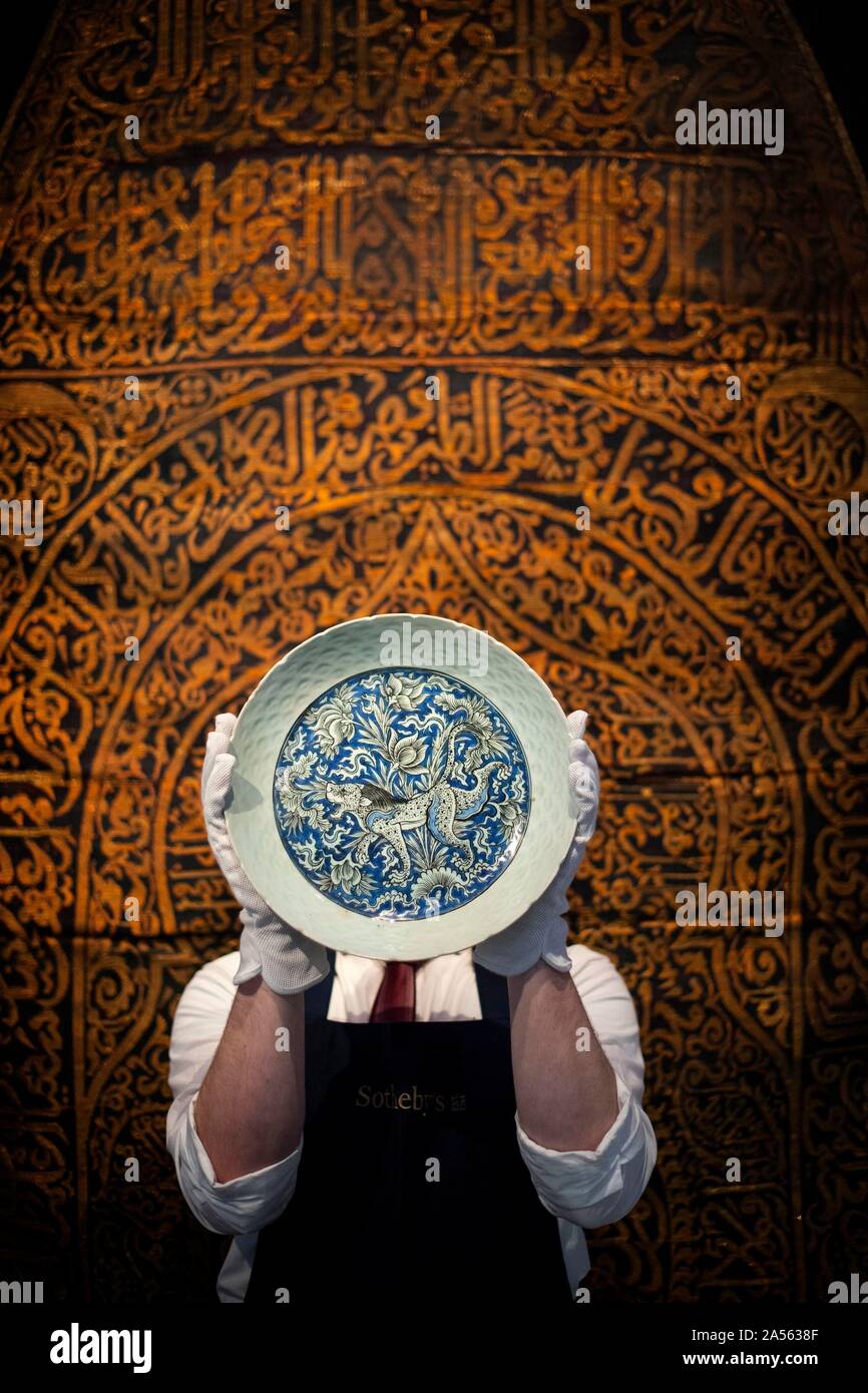 A staff member holds a Safavid pottery dish depicting a lion, made in Persia in 17th century (est. £8,000-12,000) in front of a rare Maghribi gilt-metal thread embroidered curtain, made in North Africa in 17th century (est. £50,000-70,000) during the preview of Middle Eastern Art Week at Sotheby's in London, ahead of their sale by auction on 22 and 23 October. PA Photo. Picture date: Friday October 18, 2019. Highlights of the sale will include a sumptuous leaf from the mythic 9th/10th century blue & gold Qu'ran, a dynamic painting of a great Indian epic wedding procession, jewellery including Stock Photo