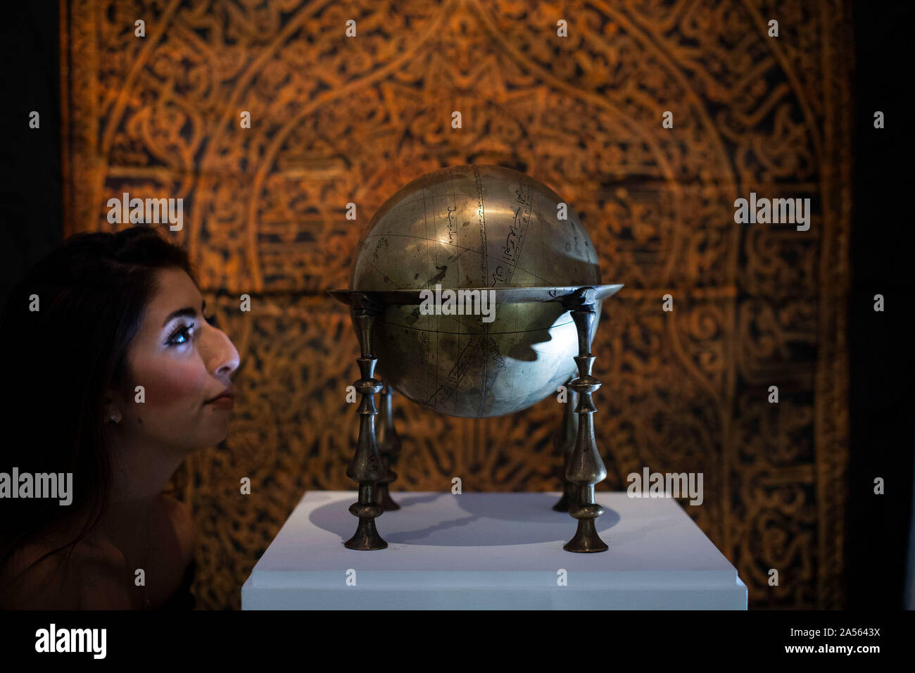 A staff member looks at a Safavid brass celestrial globe, made in Persia in 17th century (est. £80,000-120,000) in front of a rare Maghribi gilt-metal thread embroidered curtain, made in North Africa in 17th century (est. £50,000-70,000) during the preview of Middle Eastern Art Week at Sotheby's in London, ahead of their sale by auction on 22 and 23 October. PA Photo. Picture date: Friday October 18, 2019. Highlights of the sale will include a sumptuous leaf from the mythic 9th/10th century blue & gold Qu'ran, a dynamic painting of a great Indian epic wedding procession, jewellery including a Stock Photo