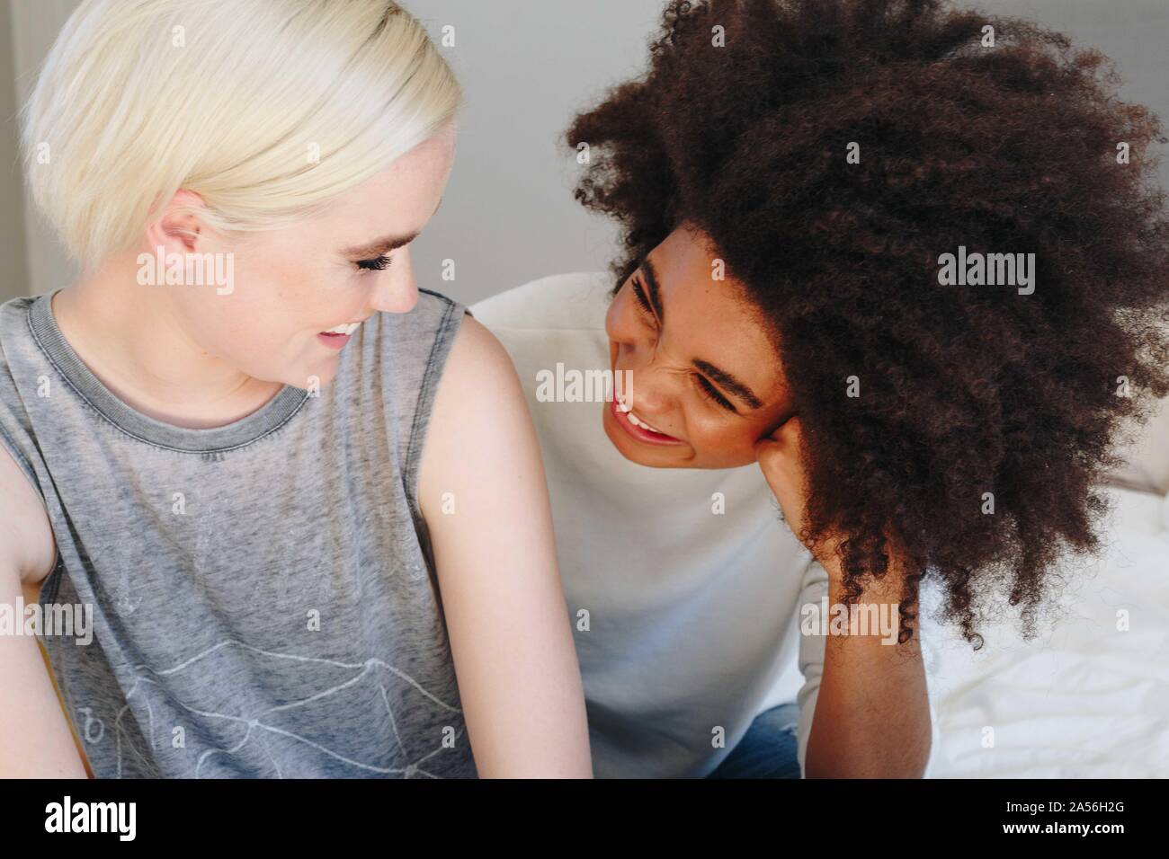 Two happy young female friends sitting on bed chatting Stock Photo