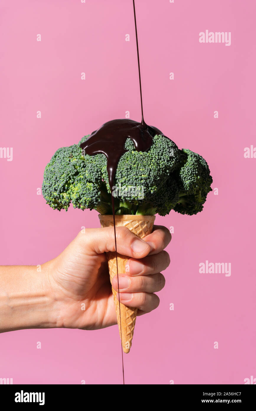 Studio shot of man's hand holding ice cream cone with broccoli on top and pouring chocolate sauce, against pink background Stock Photo