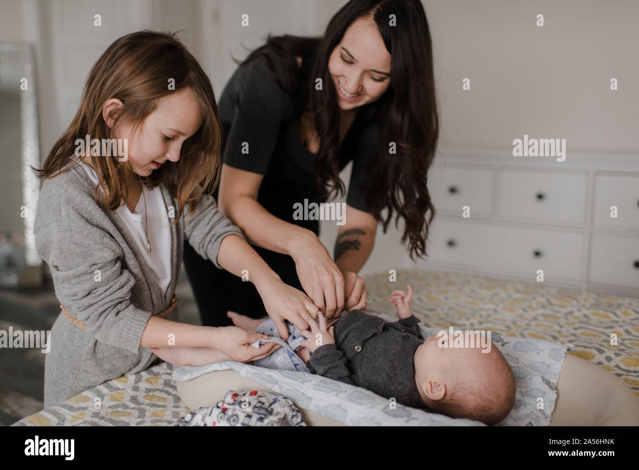 Girl helping mother change baby brother's diaper on bed Stock Photo