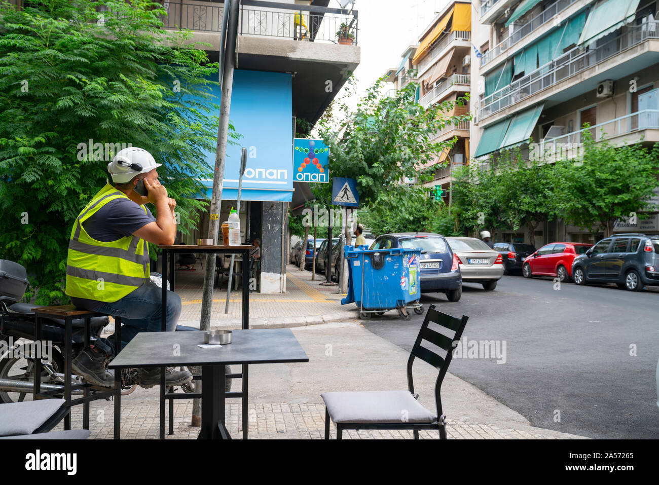 Athens Greece July 17 2019; Man sitting on stool outside cafe wearing high visibility vest, hard hat and holding mobile phone to his ear. Stock Photo