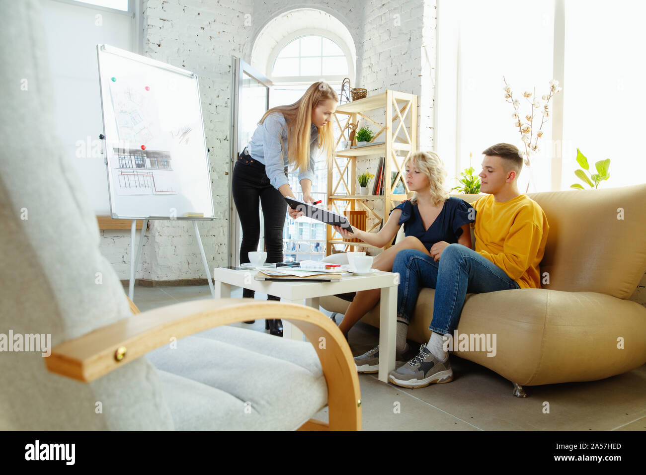 Interior designer working with young couple. Lovely family and professional designer or architector discussing conept of future interior, working with colour palette, room drawings in modern office. Stock Photo