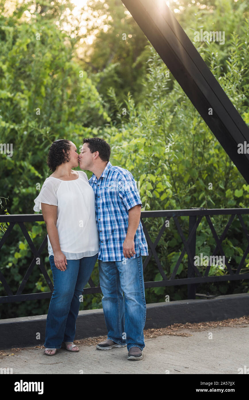 40-yr-old couple kissing on steel bridge amid lush foliage and sunligh Stock Photo