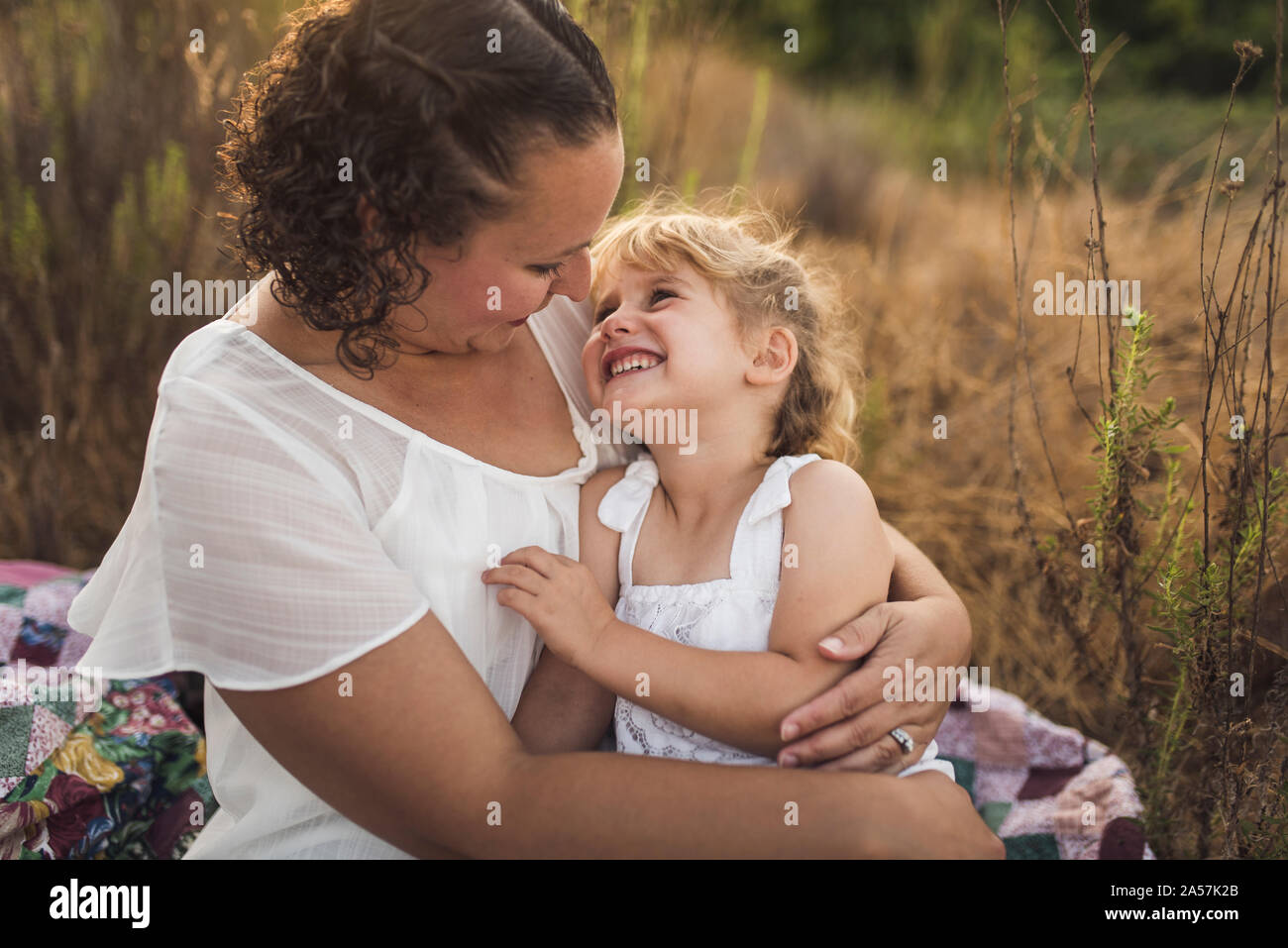 Dark-haired mom cuddles smiling young daughter outdoors on quilt Stock Photo