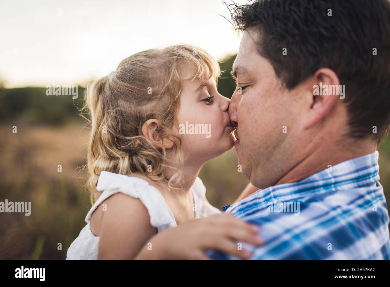 Silly young girl kissing her dad's nose outdoors in meadow Stock Photo