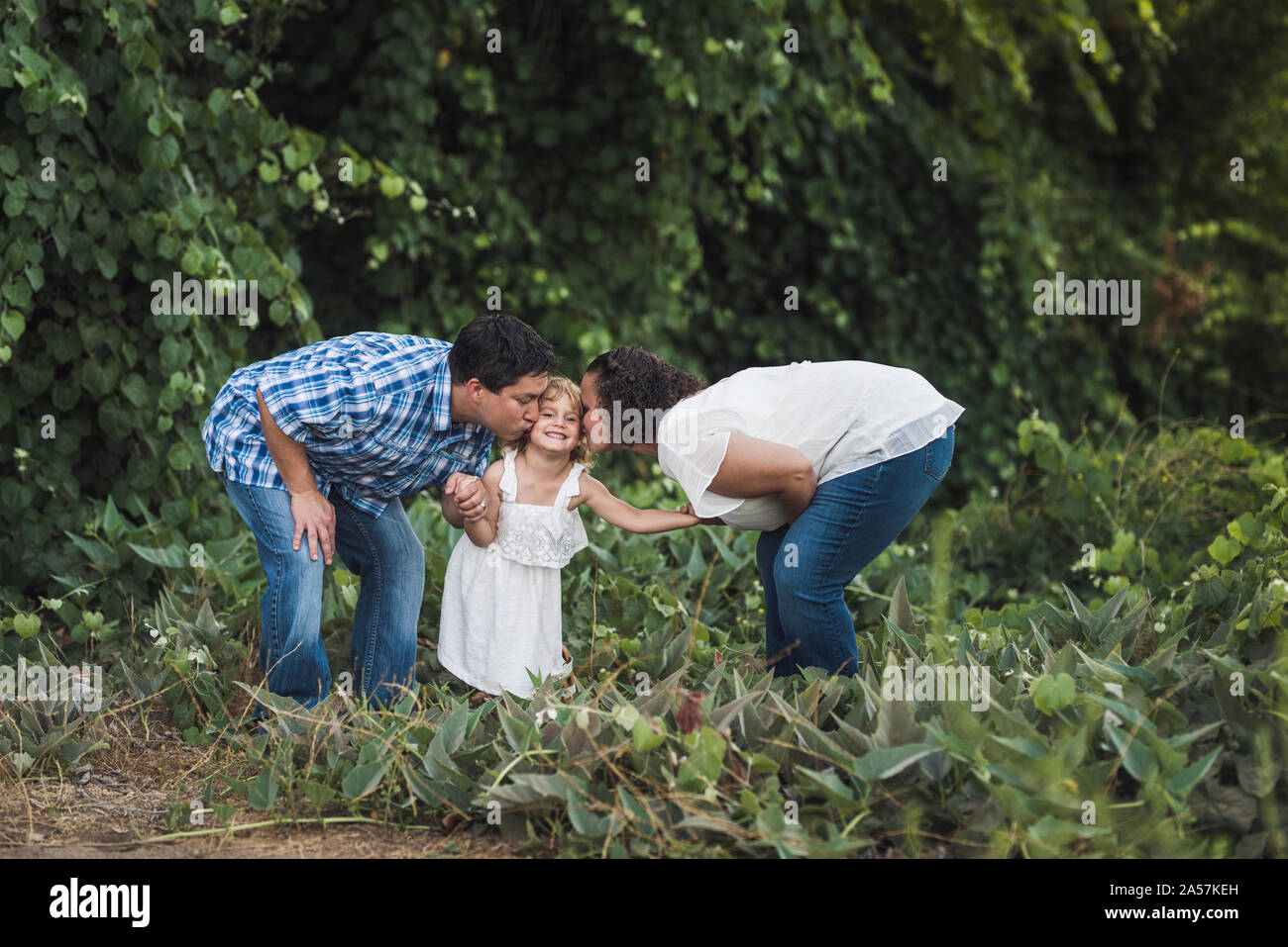 Mom & dad bending down to kiss young daughter near wall of vegetation Stock Photo