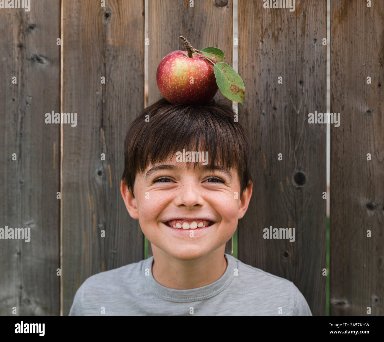 Boy smiling while balancing an apple on his head against wooden fence Stock Photo